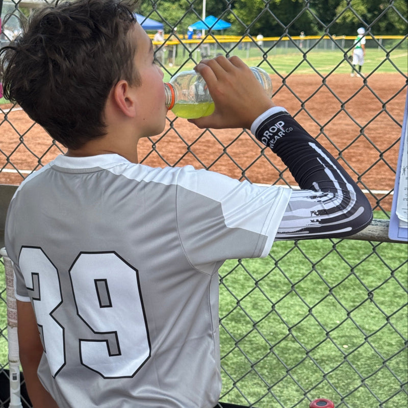 Person in a sports uniform and drip design arm sleeve standing in front of a fence with sports equipment.