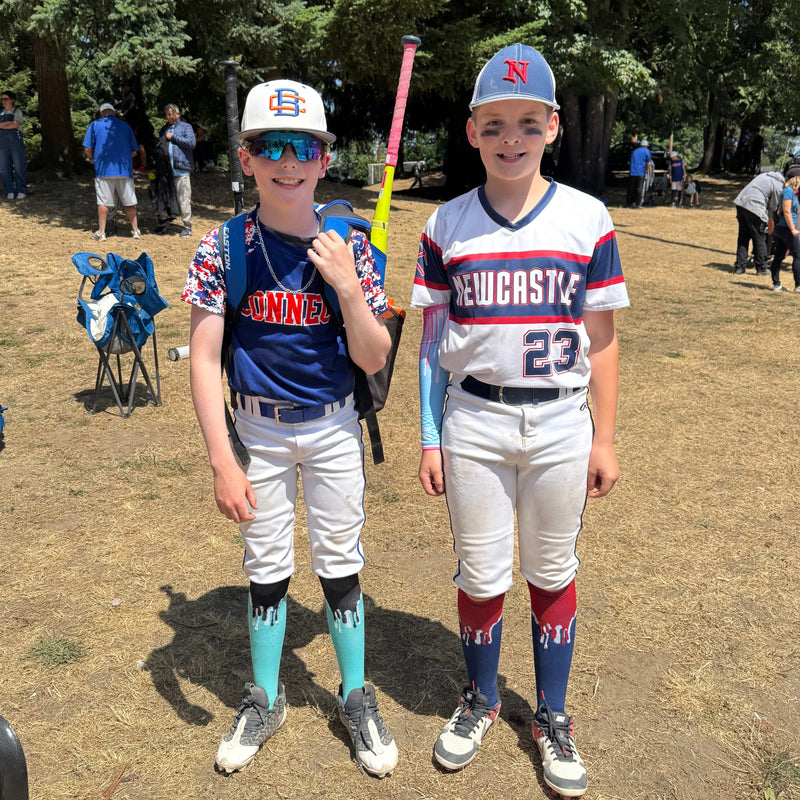 Two boys wearing baseball uniforms and DropSox standing on a dirt field with trees in the background